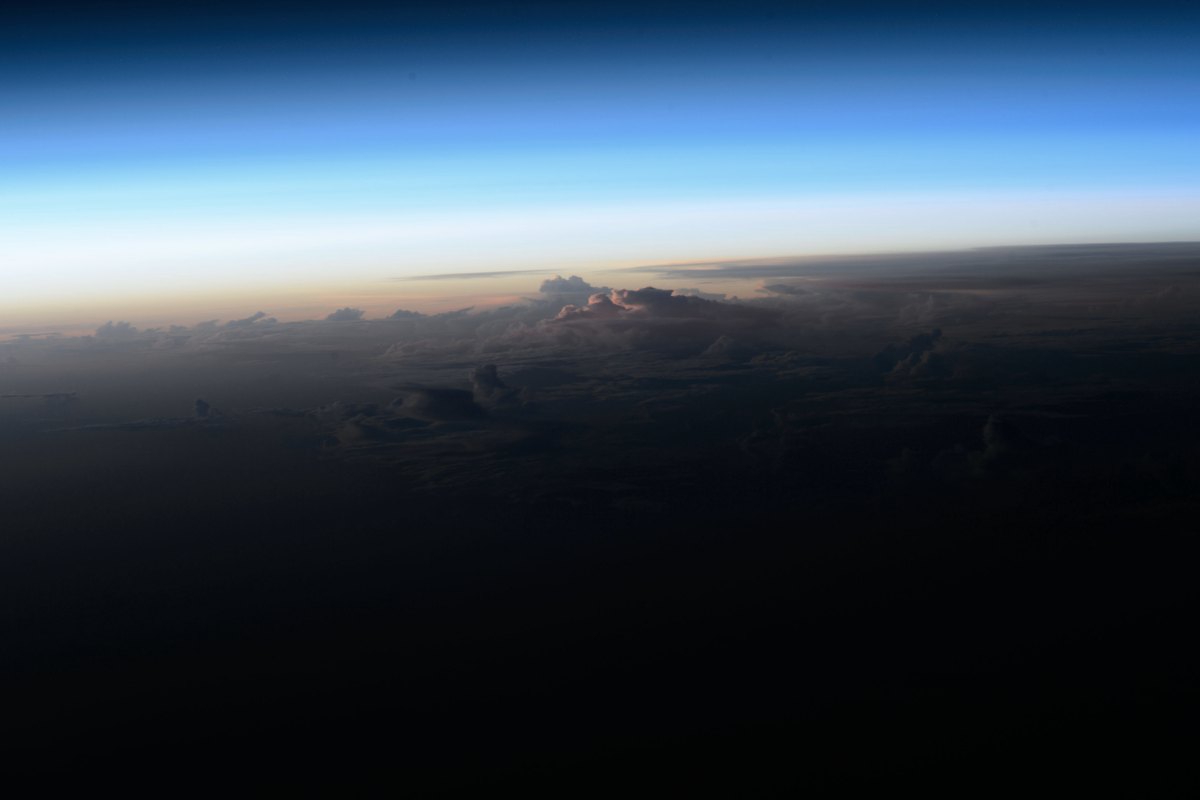 Blue and cream bands of Earth's atmosphere above a dark cloud-covered horizon at sunrise, seen from orbit
